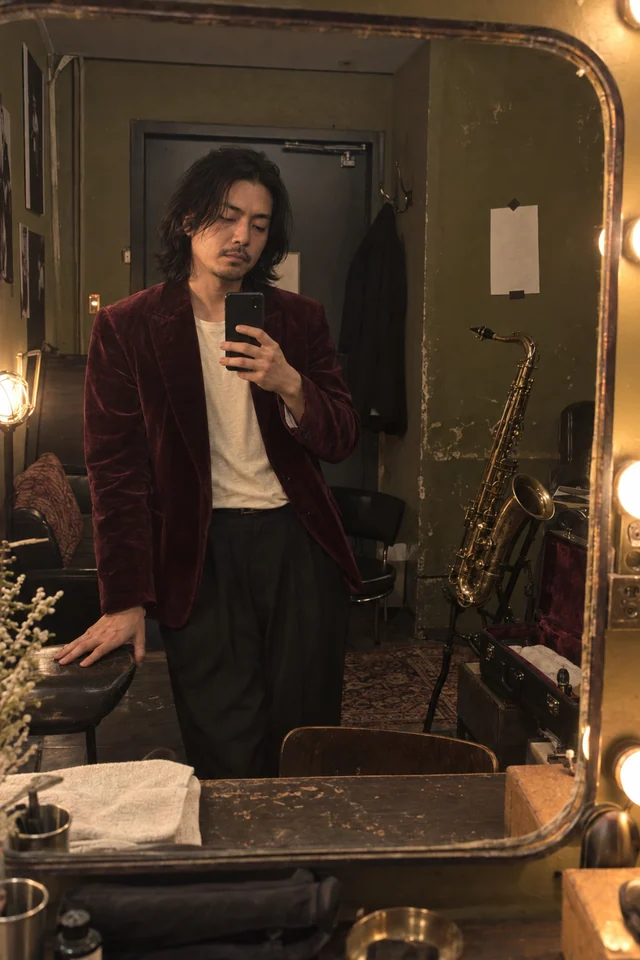 A jazz saxophonist in a burgundy jacket takes a casual phone photo in a warm backstage dressing-room mirror.